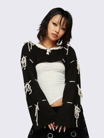 Black knitted shrug with white tie bows, designed in a bolero style with long, wide sleeves.