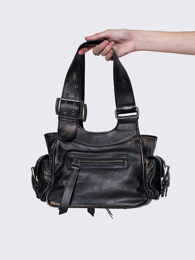Black leather handbag with multiple pockets, buckles, and a star design, held by a hand, featuring an edgy, utilitarian style.