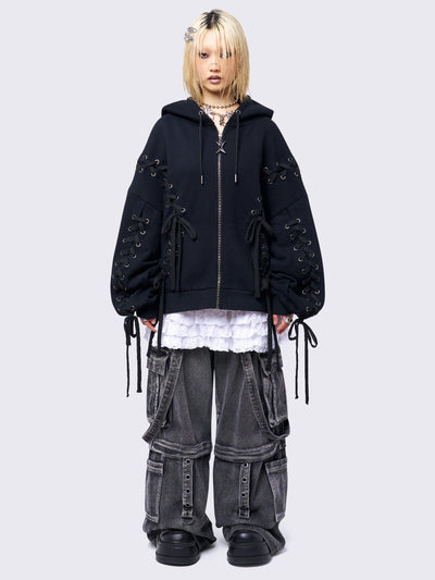 Black zip-up hoodie with lace corset details along the sleeves and front. Features a drawstring hood, zip fastening, long sleeves, and an oversized fit.