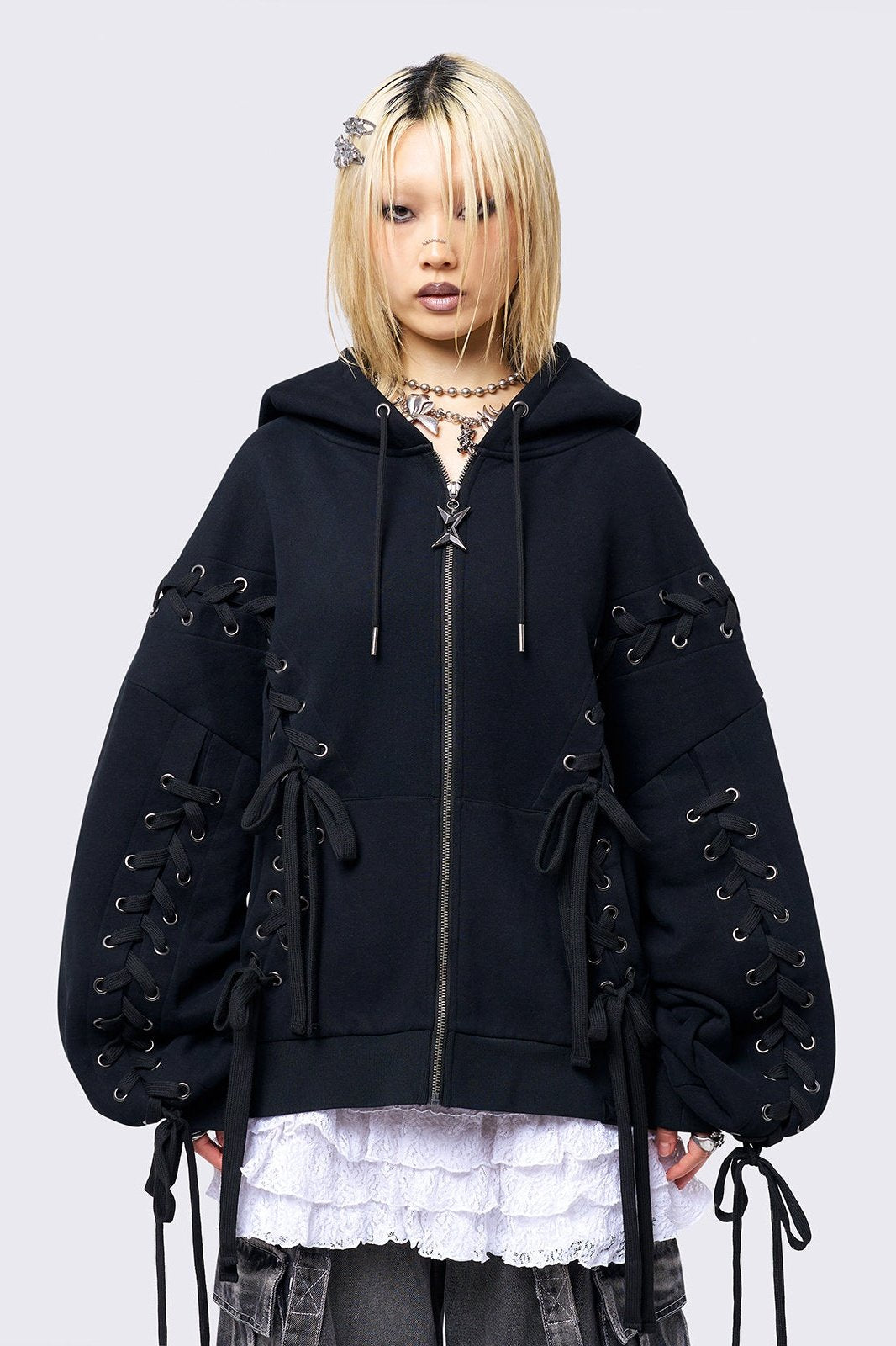 Black zip-up hoodie with lace corset details along the sleeves and front. Features a drawstring hood, zip fastening, long sleeves, and an oversized fit.