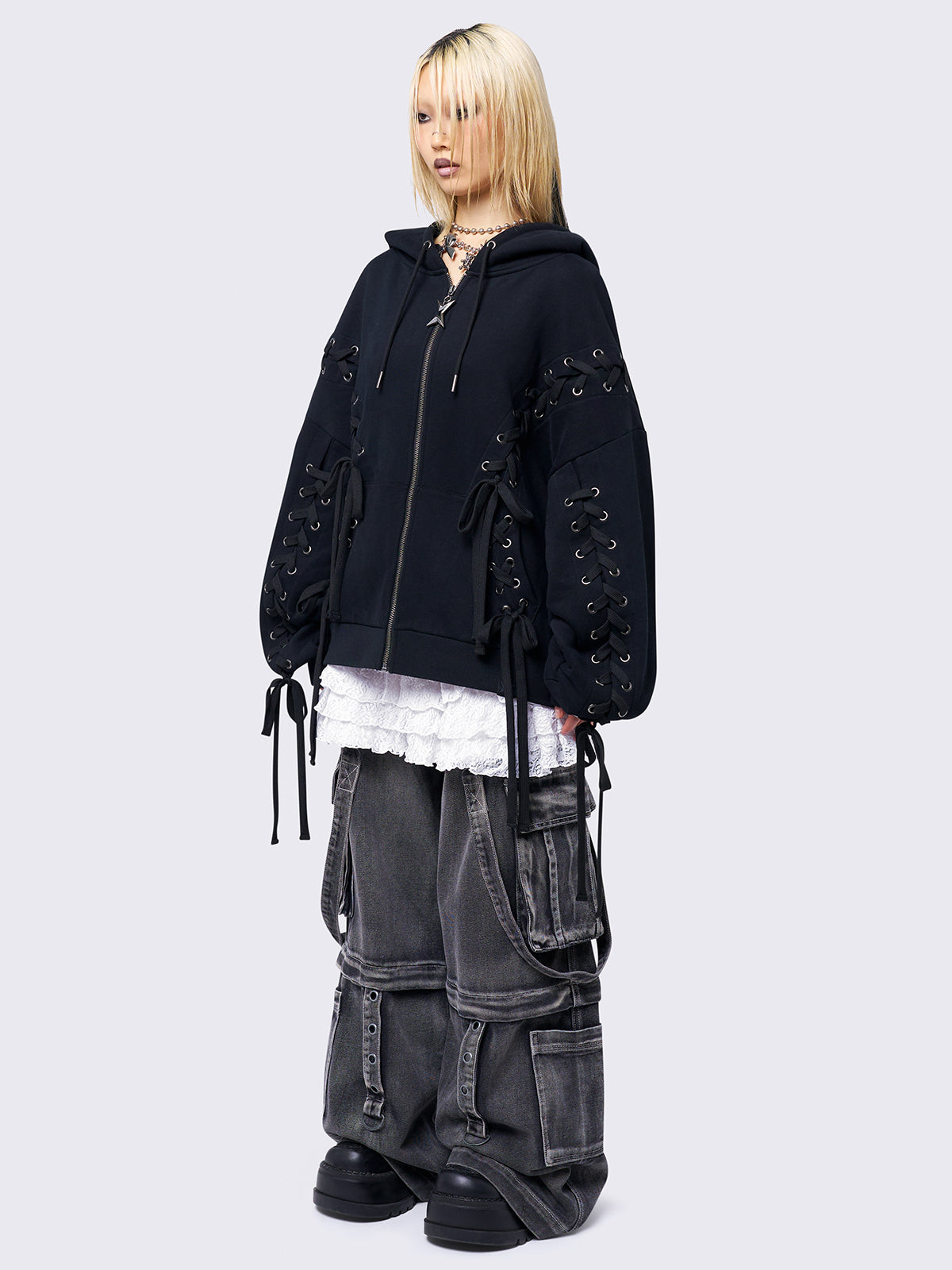 Black zip-up hoodie with lace corset details along the sleeves and front. Features a drawstring hood, zip fastening, long sleeves, and an oversized fit.