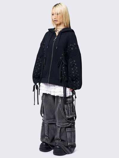 Black zip-up hoodie with lace corset details along the sleeves and front. Features a drawstring hood, zip fastening, long sleeves, and an oversized fit.
