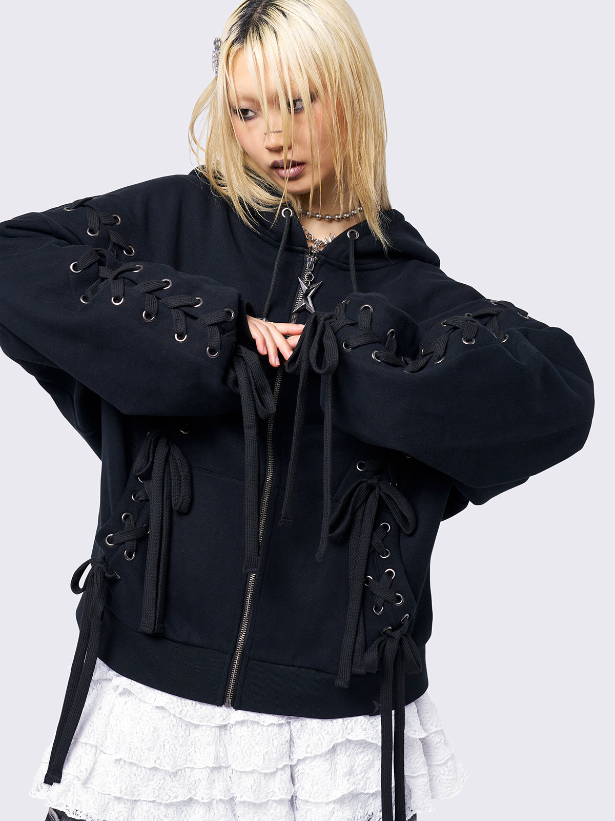 Black zip-up hoodie with lace corset details along the sleeves and front. Features a drawstring hood, zip fastening, long sleeves, and an oversized fit.