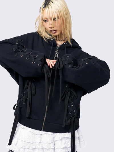 Black zip-up hoodie with lace corset details along the sleeves and front. Features a drawstring hood, zip fastening, long sleeves, and an oversized fit.
