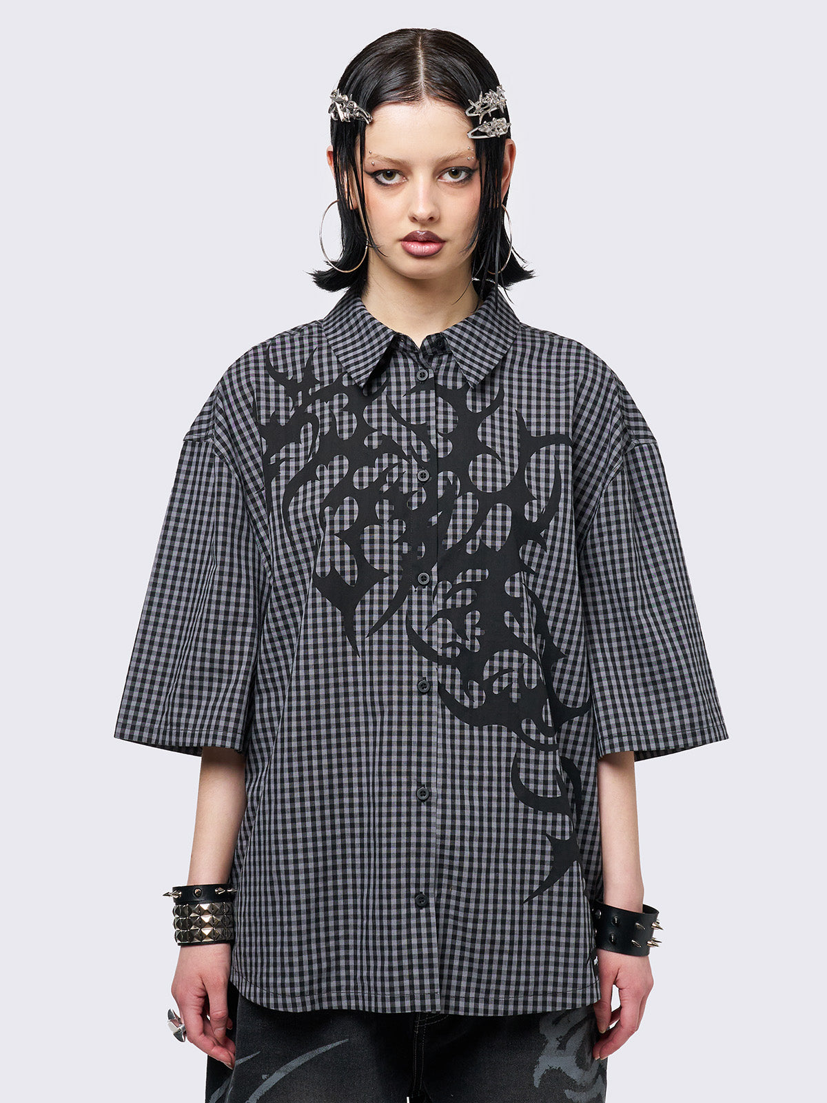 Oversized grunge-style button-up shirt with a dark grey and black checkered pattern and bold black abstract tribal graphic print. The short-sleeve design gives it a relaxed fit, while the edgy pattern adds a rebellious, alternative touch. Styled with studded bracelets for a punk-inspired look.