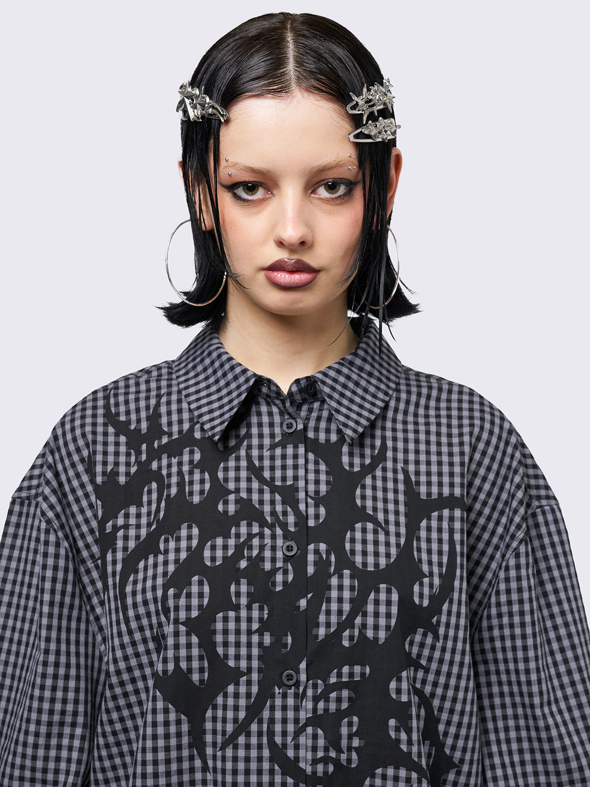 Oversized grunge-style button-up shirt with a dark grey and black checkered pattern and bold black abstract tribal graphic print. The short-sleeve design gives it a relaxed fit, while the edgy pattern adds a rebellious, alternative touch. Styled with studded bracelets for a punk-inspired look.