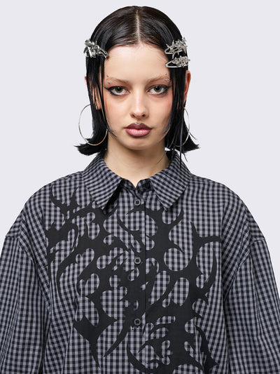 Oversized grunge-style button-up shirt with a dark grey and black checkered pattern and bold black abstract tribal graphic print. The short-sleeve design gives it a relaxed fit, while the edgy pattern adds a rebellious, alternative touch. Styled with studded bracelets for a punk-inspired look.