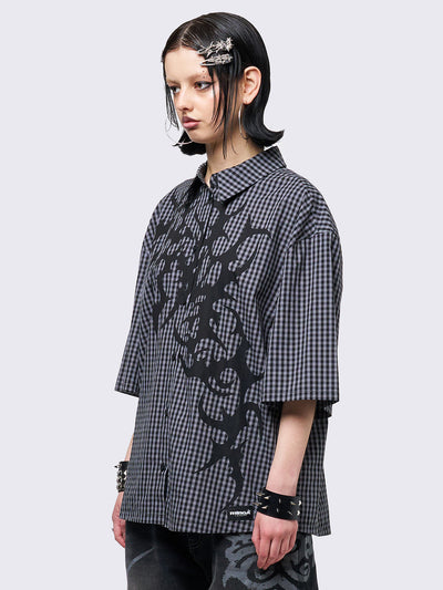 Oversized grunge-style button-up shirt with a dark grey and black checkered pattern and bold black abstract tribal graphic print. The short-sleeve design gives it a relaxed fit, while the edgy pattern adds a rebellious, alternative touch. Styled with studded bracelets for a punk-inspired look.