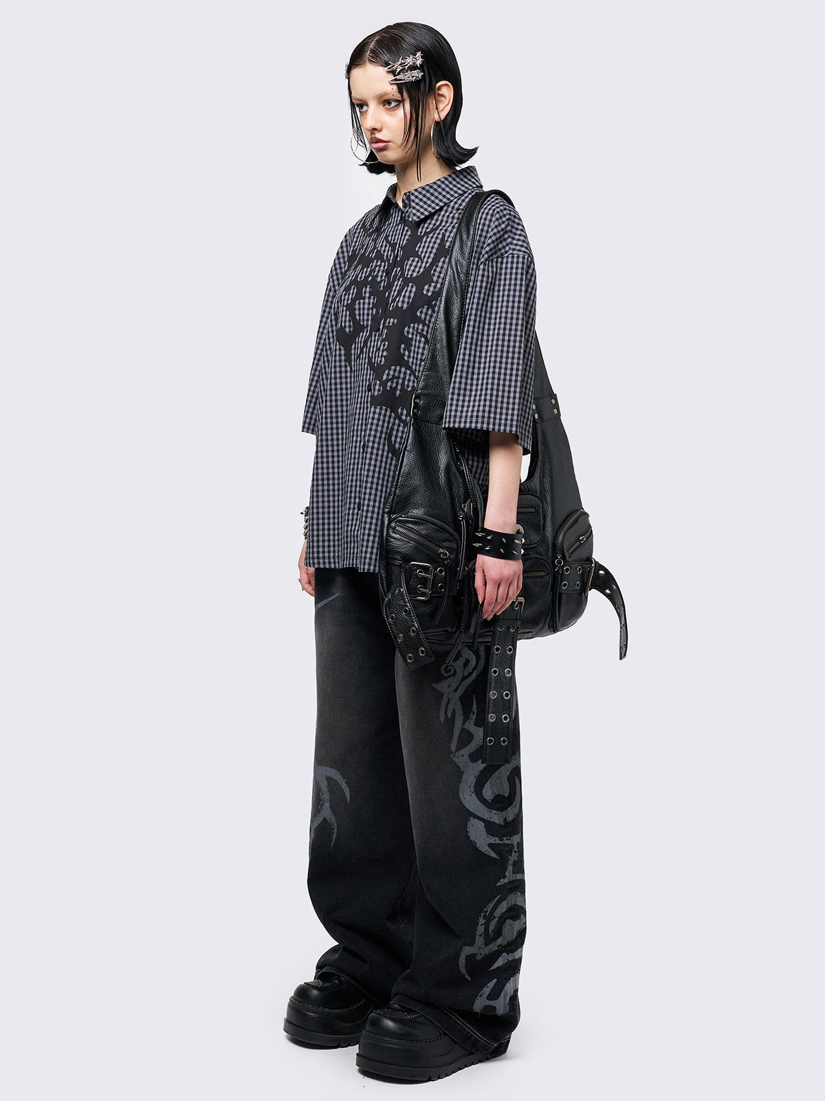 Oversized grunge-style button-up shirt with a dark grey and black checkered pattern and bold black abstract tribal graphic print. The short-sleeve design gives it a relaxed fit, while the edgy pattern adds a rebellious, alternative touch. Styled with studded bracelets for a punk-inspired look.