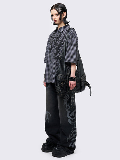 Oversized grunge-style button-up shirt with a dark grey and black checkered pattern and bold black abstract tribal graphic print. The short-sleeve design gives it a relaxed fit, while the edgy pattern adds a rebellious, alternative touch. Styled with studded bracelets for a punk-inspired look.