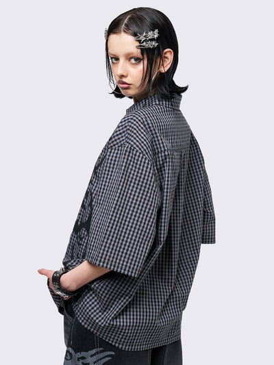 Oversized grunge-style button-up shirt with a dark grey and black checkered pattern and bold black abstract tribal graphic print. The short-sleeve design gives it a relaxed fit, while the edgy pattern adds a rebellious, alternative touch. Styled with studded bracelets for a punk-inspired look.
