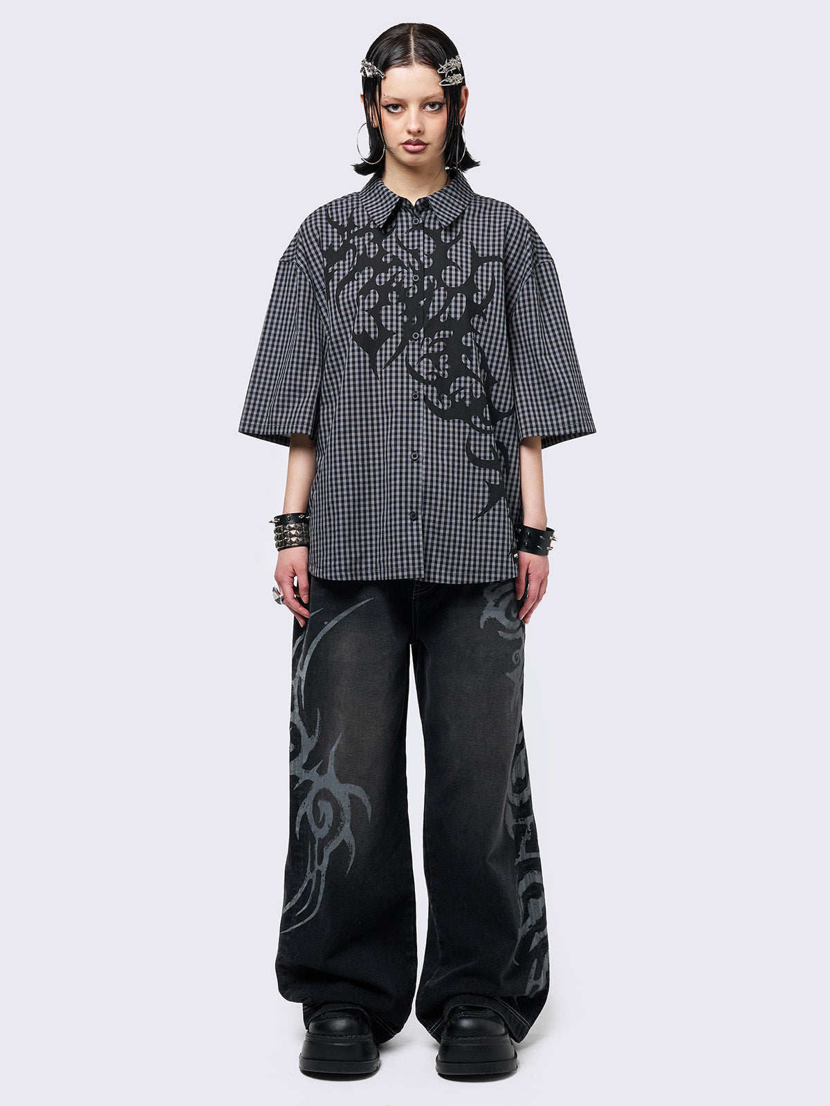Oversized grunge-style button-up shirt with a dark grey and black checkered pattern and bold black abstract tribal graphic print. The short-sleeve design gives it a relaxed fit, while the edgy pattern adds a rebellious, alternative touch. Styled with studded bracelets for a punk-inspired look.