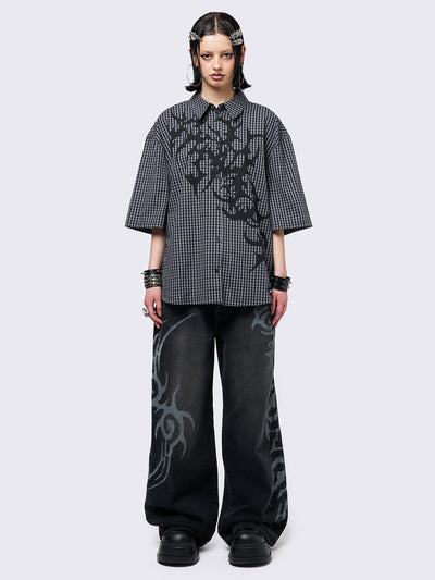 Oversized grunge-style button-up shirt with a dark grey and black checkered pattern and bold black abstract tribal graphic print. The short-sleeve design gives it a relaxed fit, while the edgy pattern adds a rebellious, alternative touch. Styled with studded bracelets for a punk-inspired look.