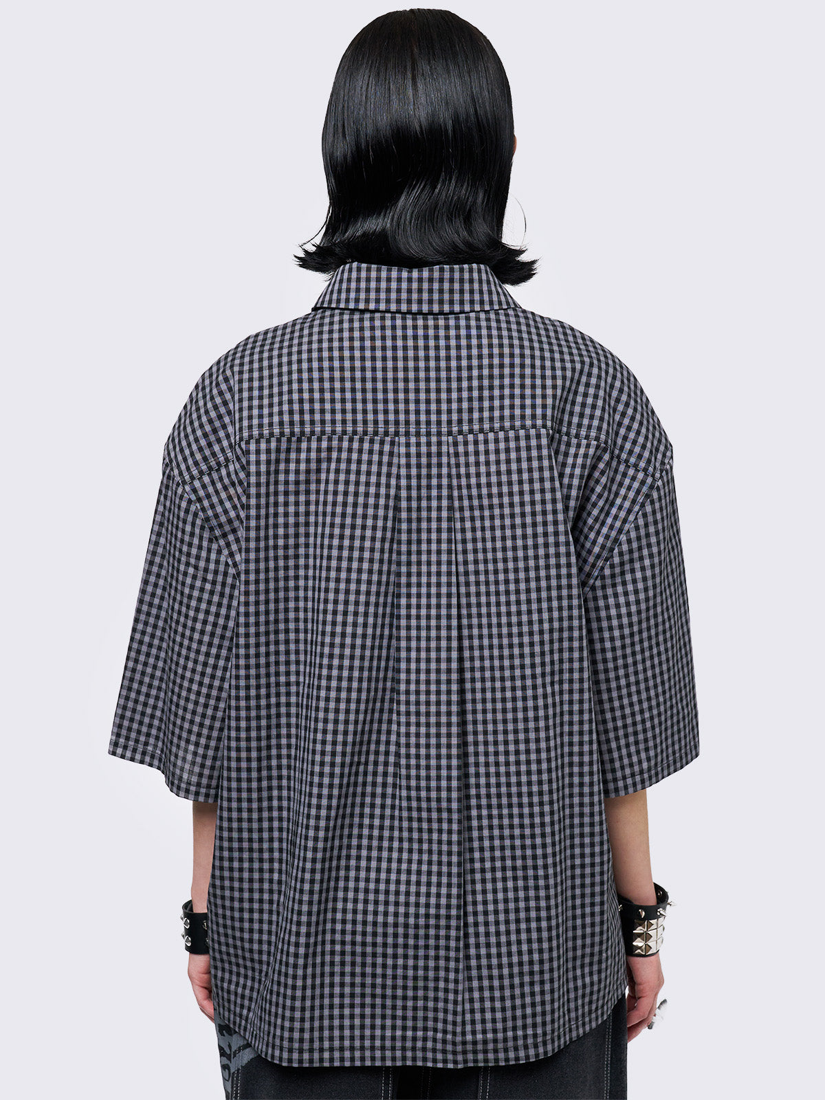 Oversized grunge-style button-up shirt with a dark grey and black checkered pattern and bold black abstract tribal graphic print. The short-sleeve design gives it a relaxed fit, while the edgy pattern adds a rebellious, alternative touch. Styled with studded bracelets for a punk-inspired look.