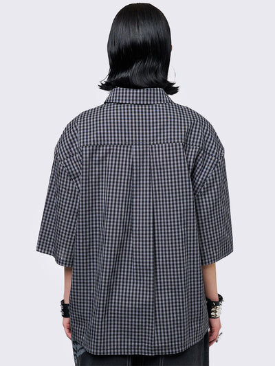 Oversized grunge-style button-up shirt with a dark grey and black checkered pattern and bold black abstract tribal graphic print. The short-sleeve design gives it a relaxed fit, while the edgy pattern adds a rebellious, alternative touch. Styled with studded bracelets for a punk-inspired look.