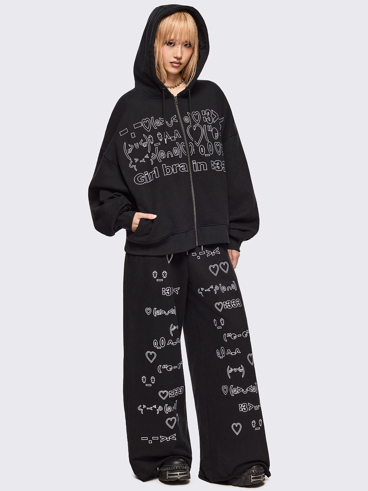 Black oversized sweatpants with all-over white emoticon and heart graphics in a playful ASCII/kaomoji style, styled in a relaxed streetwear fit.