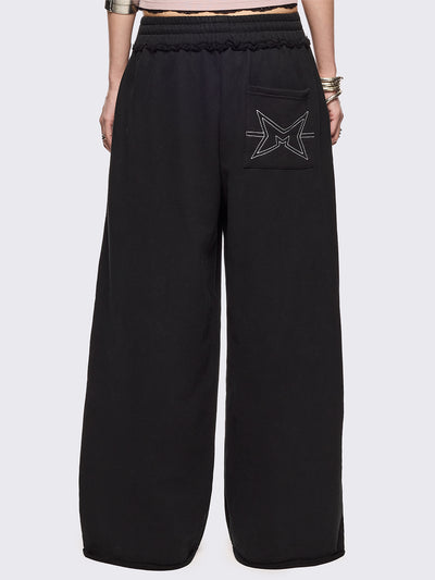 Black oversized sweatpants with all-over white emoticon and heart graphics in a playful ASCII/kaomoji style, styled in a relaxed streetwear fit.