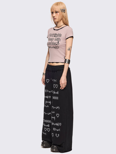 Black oversized sweatpants with all-over white emoticon and heart graphics in a playful ASCII/kaomoji style, styled in a relaxed streetwear fit.