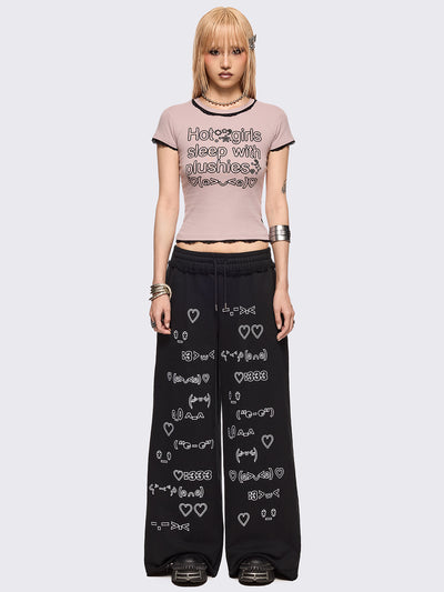 Black oversized sweatpants with all-over white emoticon and heart graphics in a playful ASCII/kaomoji style, styled in a relaxed streetwear fit.