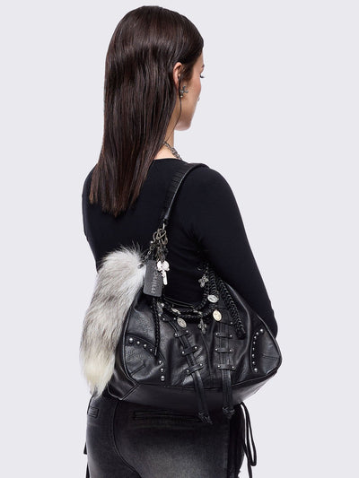 A black leather hobo bag decorated with silver studs, metallic charms, braided leather, and a large grey and white fur tail charm.