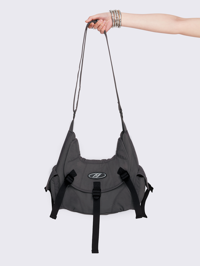 Grey nylon crossbody bag featuring multiple pockets with black webbing strap details. Flap and zip closures. Edgy, utility-inspired streetwear style.