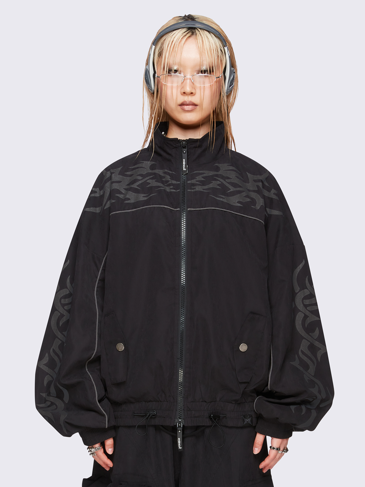 Black oversized windbreaker jacket with grey tribal graphics on the chest and sleeves, elastic cuffs, front zip closure, toggle-adjustable hem, and snap-button pockets  — edgy, utility-inspired streetwear style.